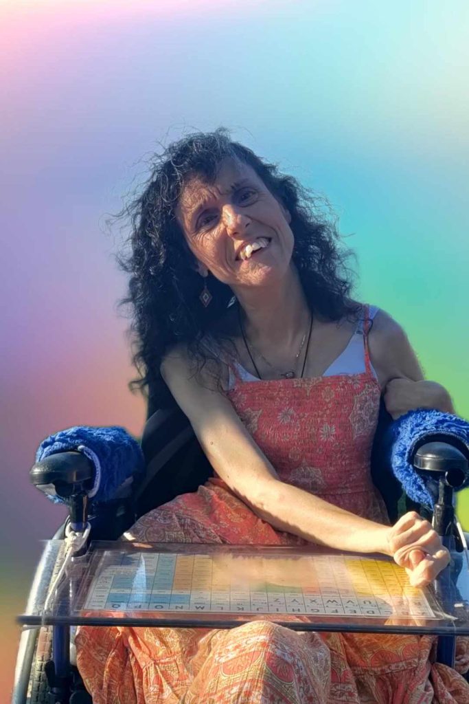 Rosermary Musachio, author of Soul of a Rose, smiling, wearing an orange flower print dress, sitting in her wheelchair with her word board.